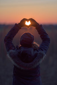 Rear view of man making heart shape against sky during sunset