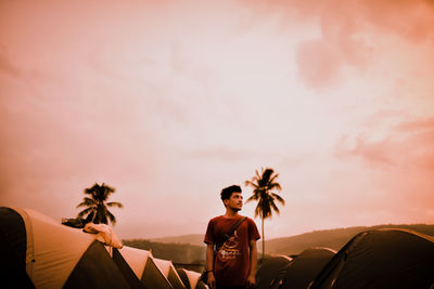 Man looking away while standing by tents against sky during sunset