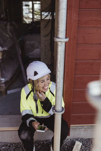 Cheerful female building contractor with smart phone sitting at construction site