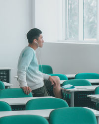 Side view of young man listening music while sitting on table in classroom