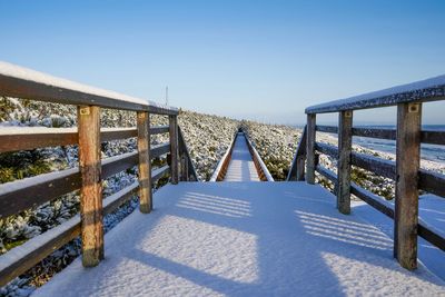 Footbridge over snow covered field against clear sky