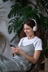 Smiling gardener woman listen music sit in armchair in indoor garden relaxed chatting in smartphone