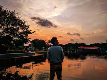 Rear view of man standing by lake against sky during sunset