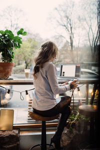 Rear view of young woman holding camera while sitting with laptop at cafe