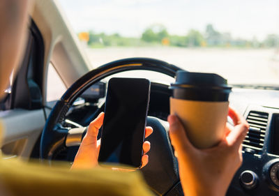 Cropped hands of woman holding smart phone and coffee in car