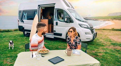 Couple sitting outdoors with camper van and beach on background