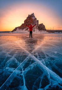 Rear view of man standing on rock by sea against sky during sunset