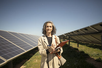 Portrait of confident senior female entrepreneur holding folder while standing near solar panels in field