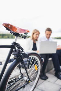 Cropped image of bicycle on sidewalk with business people working on laptop against clear sky