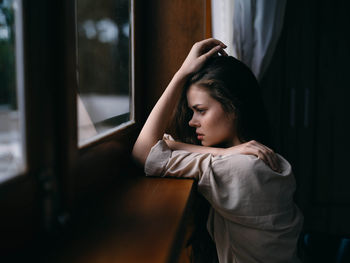 Side view of young woman looking through window at home