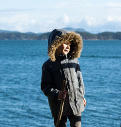 Boy wearing warm clothing holding stick while standing by sea against sky