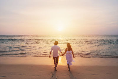 Rear view of friends enjoying at beach against sky during sunset