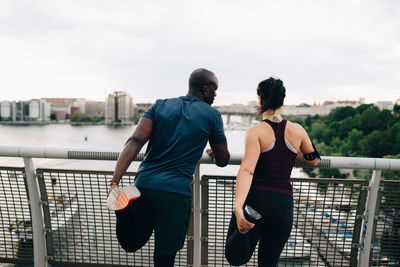 Rear view of male and female athletes stretching legs while standing on footbridge over sea in city