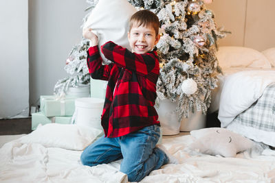 Boy in a red  shirt sits by the christmas tree with gifts on the floor and fights with a pillow