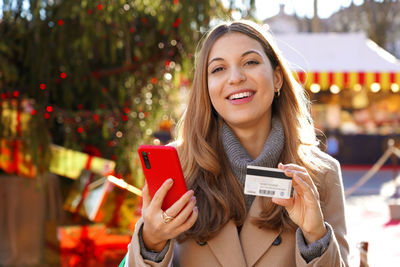 Woman holding smartphone and credit card in her hands with christmas markets on the background
