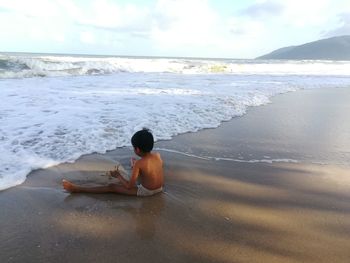 Man sitting on shore at beach against sky