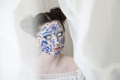 Woman with painted face and eyes closed by curtain at home