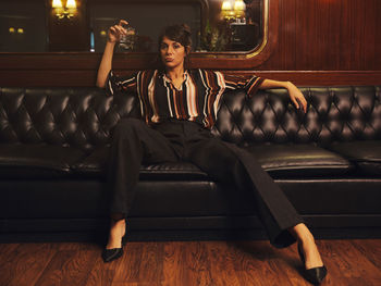 Fashionable stylish woman in trendy clothes holding glass of alcohol sitting wide spread apart legs on black leather couch and looking at camera