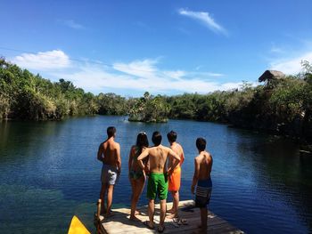 Rear view of friends standing on pier over lake against blue sky