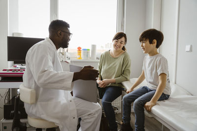 Young male pediatrician showing tablet pc to mother and son sitting in examination room at clinic