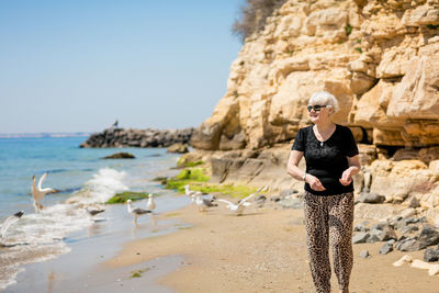 Senior woman in dark sunglasses feeds seagulls on the beach