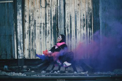 Smiling woman holding distress flare while sitting against weathered wooden wall