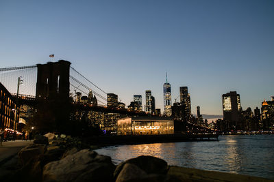 Illuminated bridge over river by buildings against clear sky