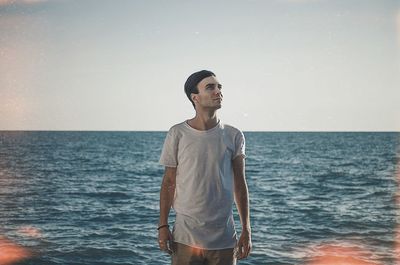 Young man looking away while standing at beach against sky