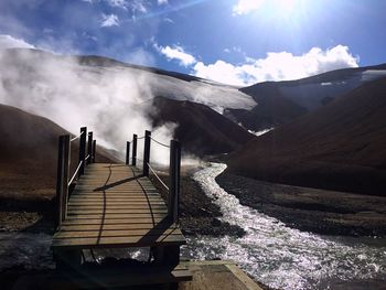 Footbridge over river against hot spring and mountains during sunny day