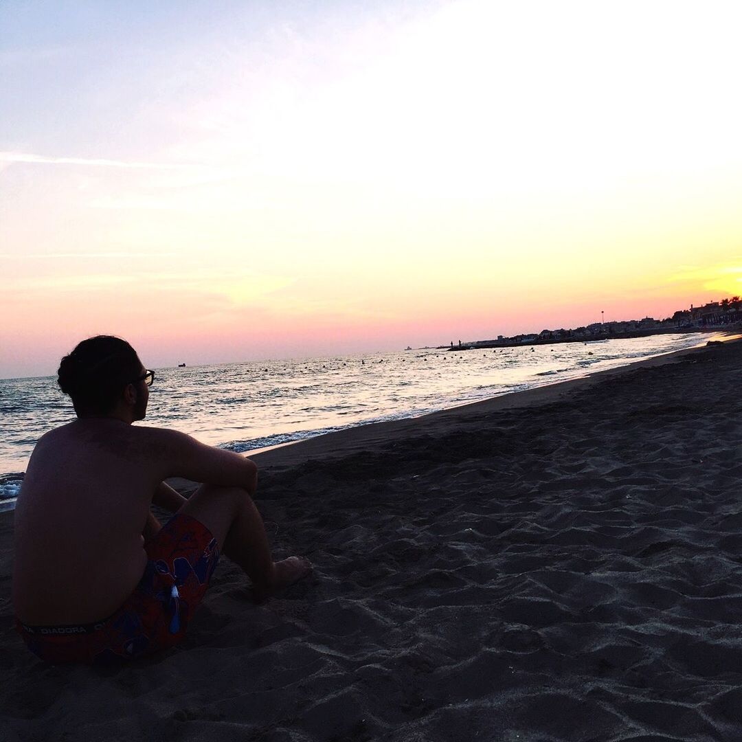 REAR VIEW OF MAN SITTING ON BEACH AGAINST SKY DURING SUNSET