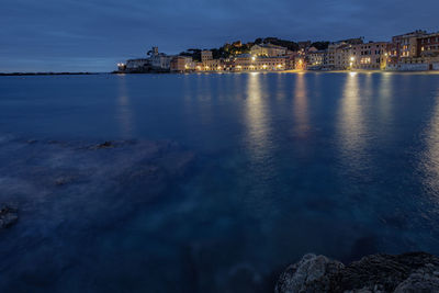 Illuminated city by sea against sky at dusk