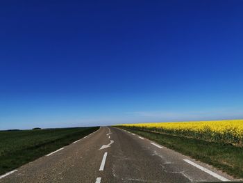 Empty road amidst field against clear blue sky