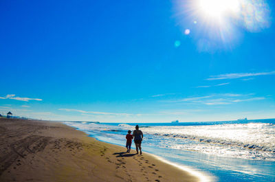 Rear view of mother and son walking at beach against blue sky during sunny day