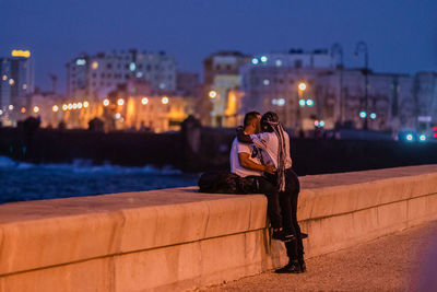 Man sitting on retaining wall against illuminated city at night