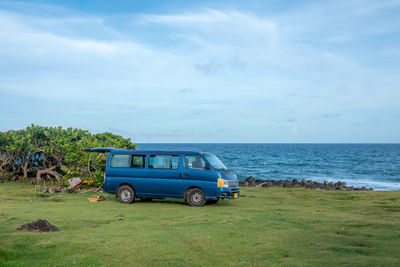 A small blue bus sitting on the cliff beside the sea.