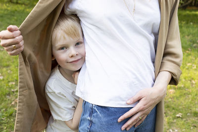 Little smiling caucasian 7 yo boy looks out mother's jacket, hiding behind her back in park. mom's