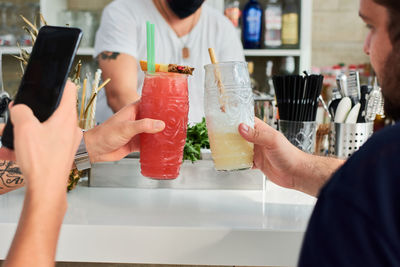 Crop unrecognizable male friends clinking glasses with fresh smoothies and taking photo on smartphone while relaxing at counter in cafe