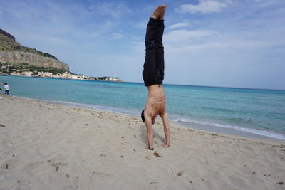 Full length of shirtless man doing handstand on sand at beach against sky