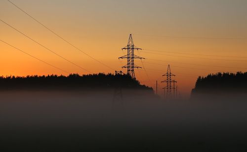 Low angle view of silhouette electricity pylons against orange sky in foggy weather