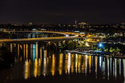 Illuminated bridge over river by buildings against sky at night