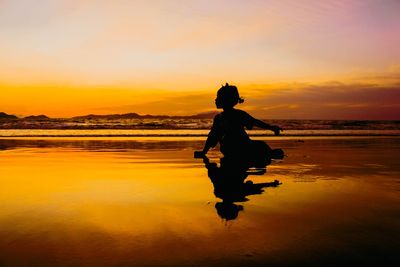 Silhouette girl sitting on beach against sky during sunset