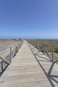 Wooden walkway leading towards sea against clear blue sky