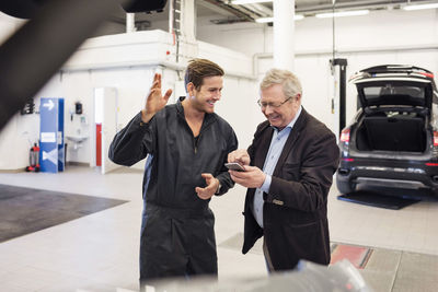 Happy mechanic with senior man using mobile app at auto repair shop