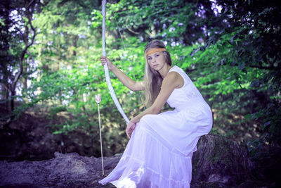 Side view of teenage girl holding bow and arrow while sitting on tree stump in forest