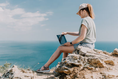Side view of woman using mobile phone while sitting on rock at beach against sky