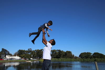Father throwing son in air by lake against clear blue sky