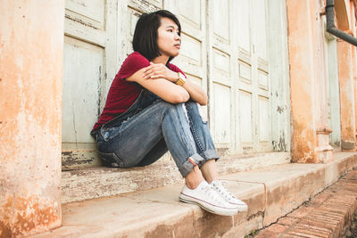 Full length of thoughtful young woman looking away while sitting against closed door