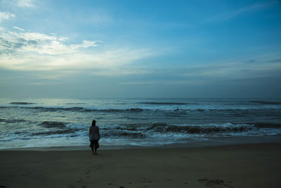 Rear view of woman standing on shore at beach against sky at dusk