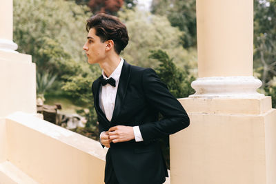 A beautiful young man, the groom in an elegant wedding suit, stands posing in the city's old park