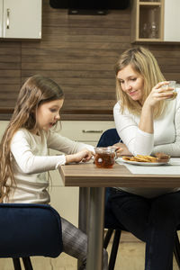 White mom and little daughter talks while having breakfast in kitchen, girl holds cell phone, device